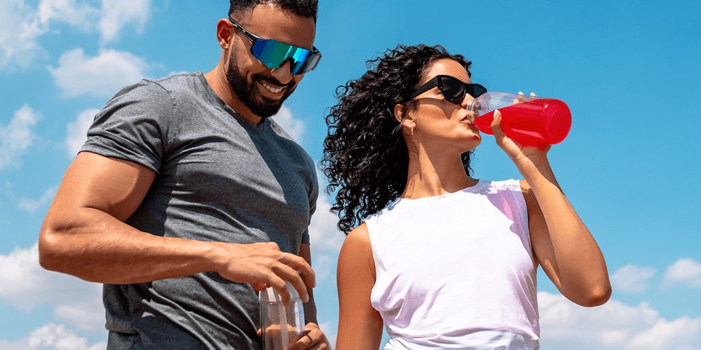 A smiling man and a woman in sunglasses drinking a red energy drink from a clear shaker bottle outdoors under a blue sky