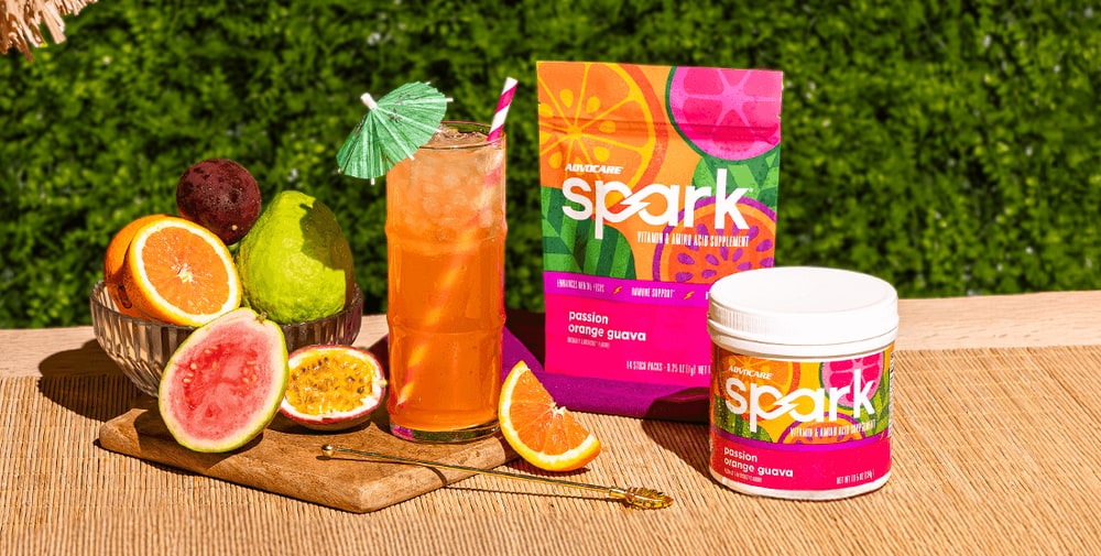 A pouch and a canister of passion orange guava AdvoCare Spark beside an iced orange drink and cut tropical fruit on a woven table