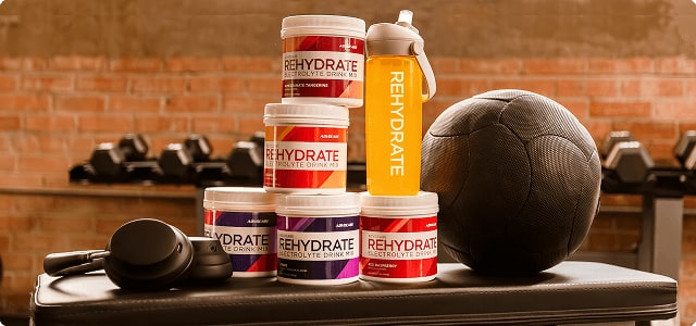 A stack of AdvoCare Rehydrate canisters and an orange drink bottle on a gym bench beside a medicine ball and headphones