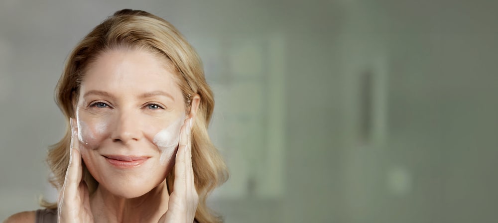 Smiling woman with blonde hair applying white Aloette cream to both cheeks in a softly lit bathroom