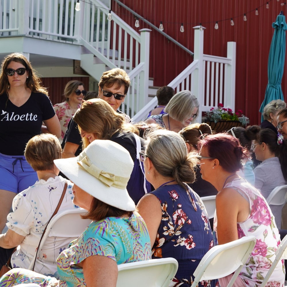 Group of Aloette consultants, one wearing an Aloette T-shirt, seated together outdoors at a company event in front of a red barn