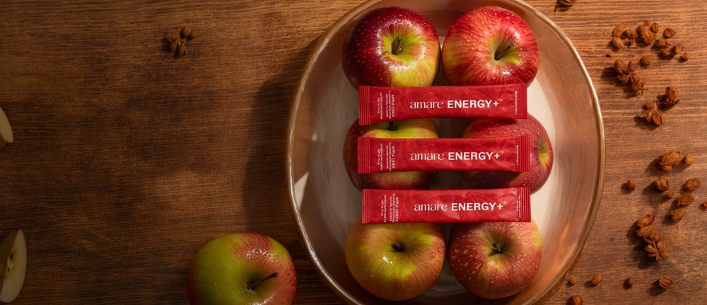 Three red Amare Energy+ Apple Cider stick packs laid across a bowl of apples on a wooden table