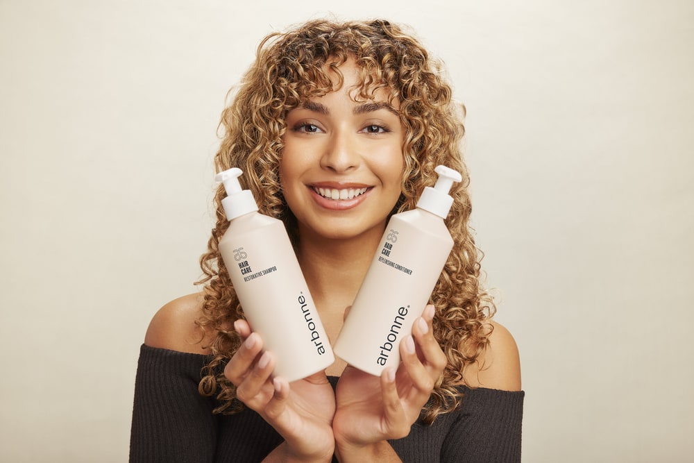 Smiling woman with curly hair holding Arbonne Restorative Shampoo and Replenishing Conditioner bottles