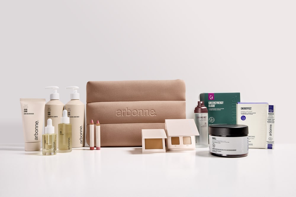 Arbonne hair care bottles, face oils, lip pencils, makeup compacts, a GreenSynergy box, BeWell jar and EnergyFizz sticks around a tan cosmetics bag