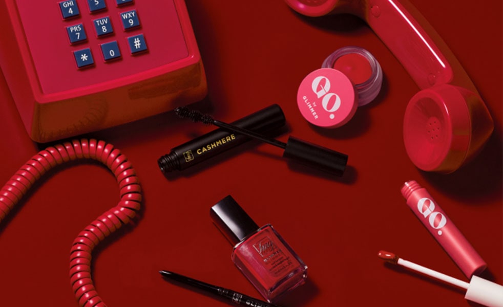 Avon cashmere mascara, Glimmer lip color, nail polish and lip gloss laid out on a red surface next to a vintage red telephone