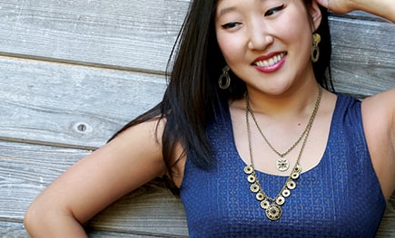 A smiling woman in a navy top wearing a layered gold coin necklace and matching earrings against a weathered wood wall