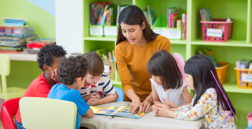 A teacher reads a picture book with five young children around a low table in a bright classroom library