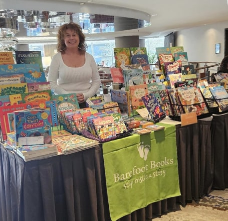 A Community Bookseller stands behind a long display of Barefoot Books picture books at an indoor event, with a green Barefoot Books table runner