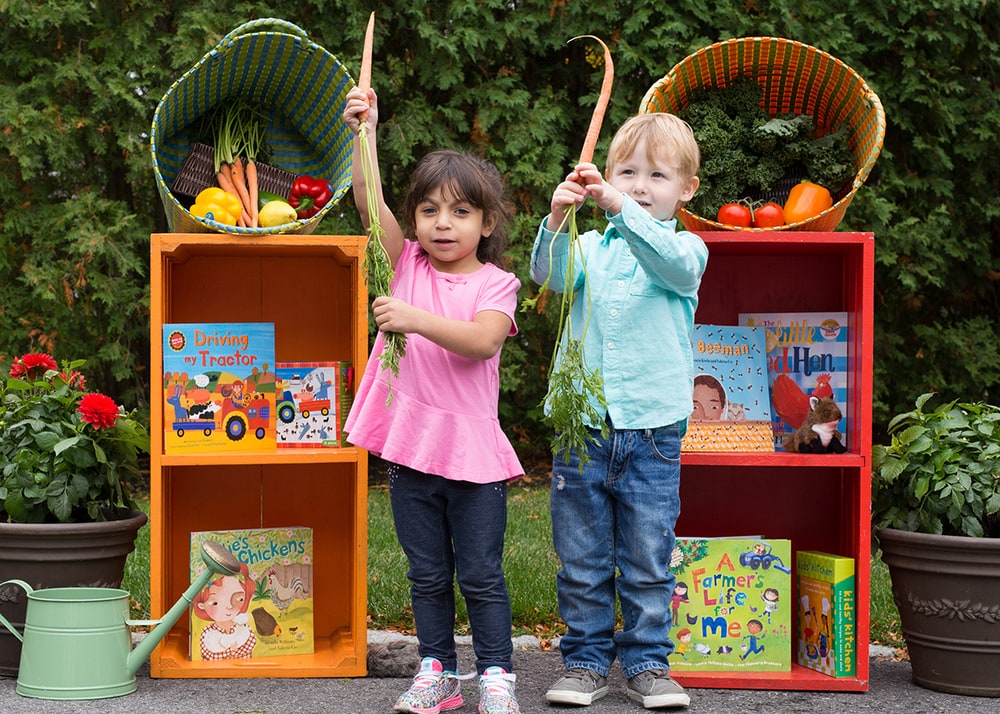 Two young children hold up fresh carrots between orange and red crates filled with Barefoot Books picture books about farms and food