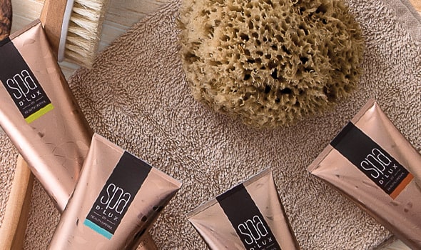 Rose-gold Spa D'Lux body care tubes laid out on a soft towel with a natural sea sponge and a wooden bath brush