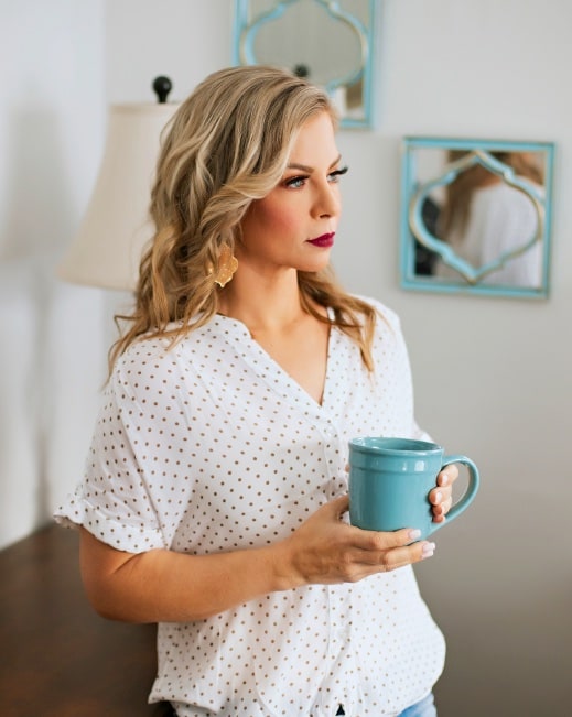 A woman with wavy blonde hair and berry lipstick holding a teal mug at home