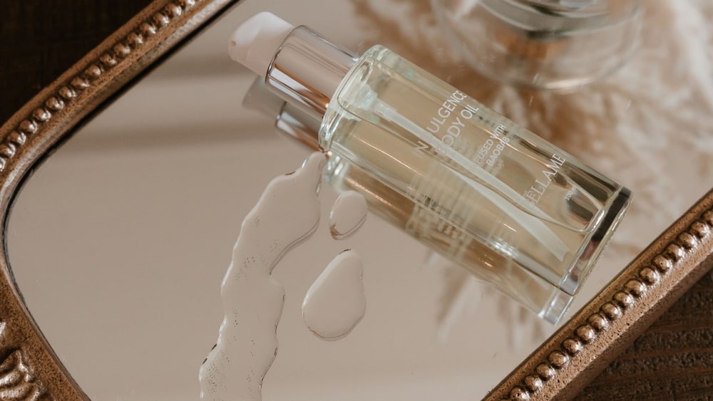 A glass bottle of Bellame Indulgence Body Oil resting on a gold-framed mirror tray beside drops of oil