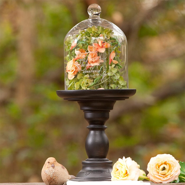 A glass cloche filled with peach flowers and greenery on a dark wooden pedestal, set outdoors beside cream roses and a small ceramic bird
