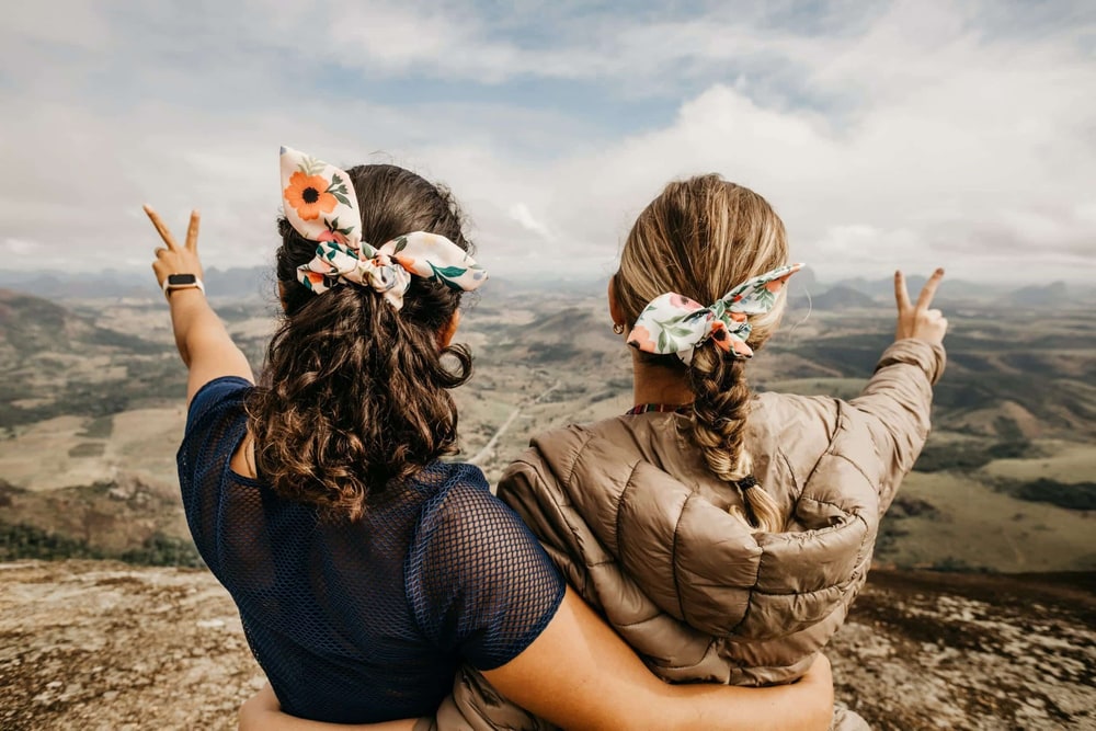 Two women with flowered hair scarves hug on a mountaintop and flash peace signs over a wide valley