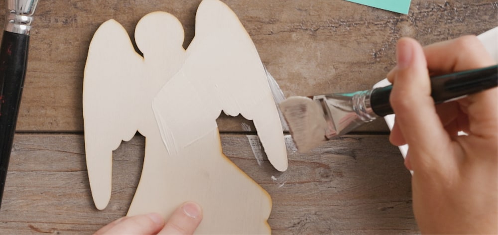 Hands brushing white paint onto a wooden angel cutout on a rustic wood table