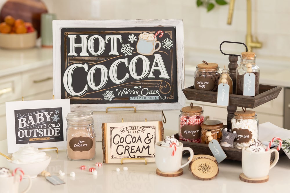 A hot cocoa bar on a kitchen counter with a framed chalkboard sign, labeled jars of toppings, mugs and a wooden sign