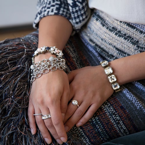 A woman's hands resting on a woven blanket, wearing several Chloe + Isabel crystal bracelets and rings