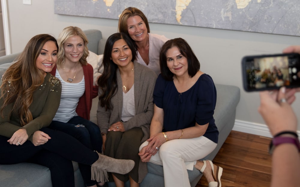 Five smiling women sitting together on a grey sofa while a friend takes their photo with a phone
