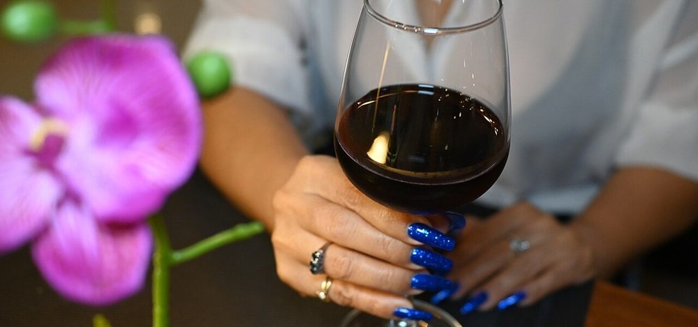 A woman with glittering blue nails holding a glass of red wine at a table beside a pink orchid, the kind of evening a home party aimed for
