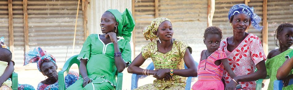 A group of women and children in bright printed dresses sitting on plastic chairs in a village, part of the artisan communities Color by Amber worked with