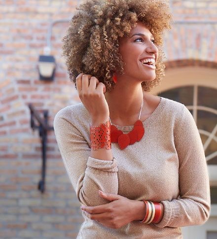 A smiling woman in a beige sweater wearing a red cut-out cuff bracelet and a red petal statement necklace