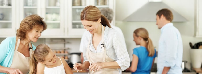 A family cooking together in a bright white kitchen, a grandmother and girl stirring a bowl beside a smiling woman in an apron