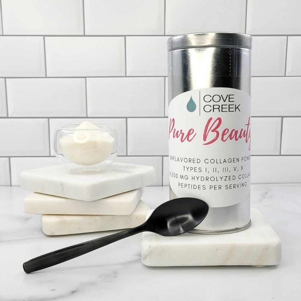 A silver tin of Cove Creek Pure Beauty collagen powder on marble coasters beside a small glass dish of powder and a black spoon