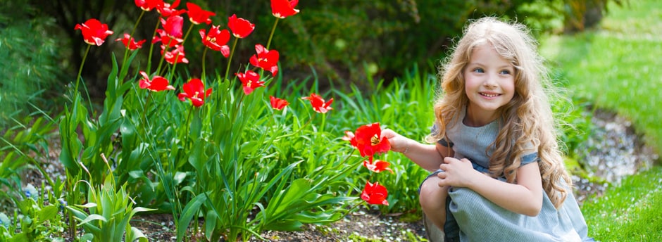 A young girl in a blue DakoModa dress crouching beside red tulips in a garden