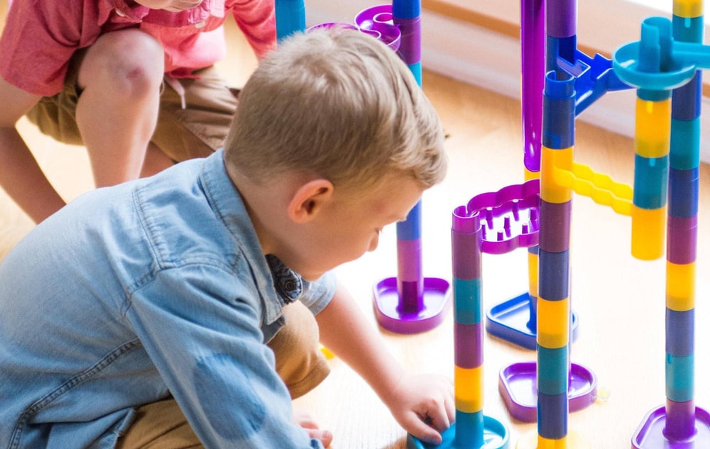 Two young boys building a tall purple, blue and yellow Marbleworks marble run on a wooden floor