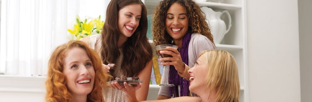 Four laughing women at a home party sharing a plate of chocolates, one holding a chocolate martini