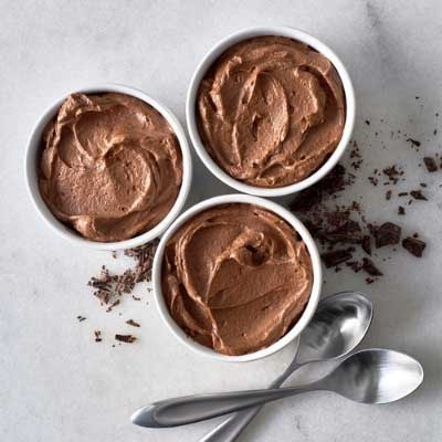 Three white ramekins of whipped chocolate mousse with spoons and chocolate shavings on a marble surface