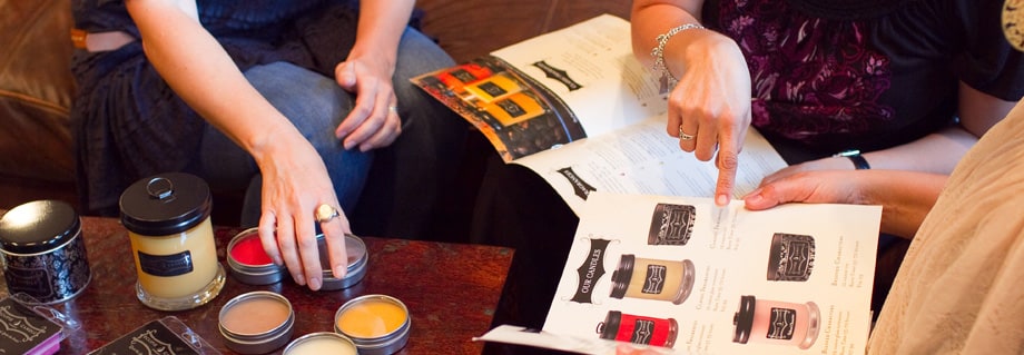 Women at a home party flipping through a candle catalog beside jar candles and open tins of wax melts on a coffee table