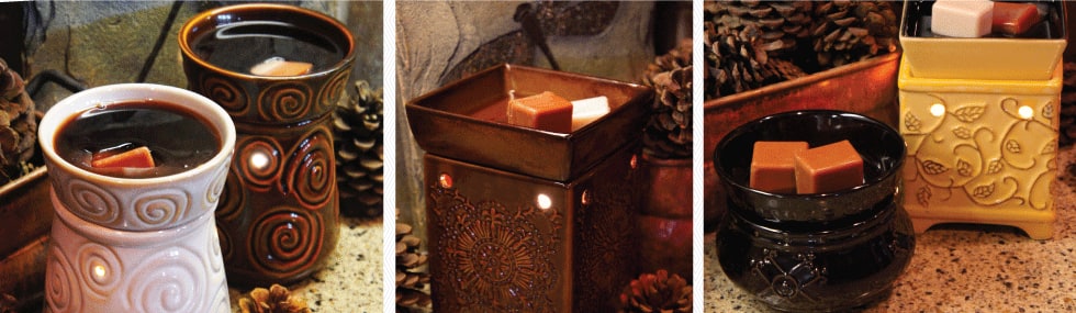 Three styles of ceramic wax warmers holding cubes of melting wax, arranged among pine cones