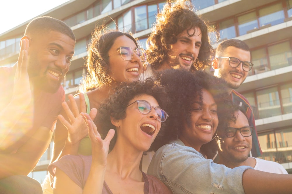 A group of smiling friends crowding together for a selfie in warm evening sunlight outside an apartment building