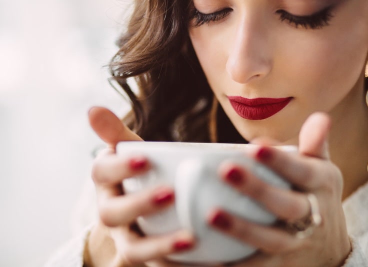 A woman with red nail polish holding a white coffee cup close to her face