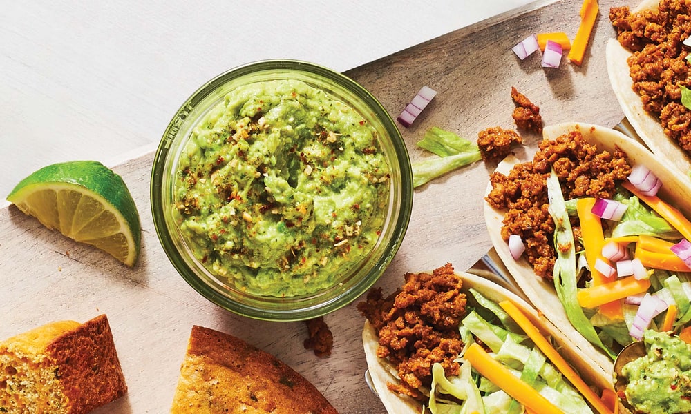 A glass jar of guacamole beside a lime wedge, cornbread and tacos filled with seasoned ground meat, cheese and lettuce