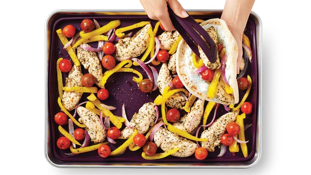 Hands lifting seasoned chicken strips, peppers and cherry tomatoes off a sheet pan into a pita