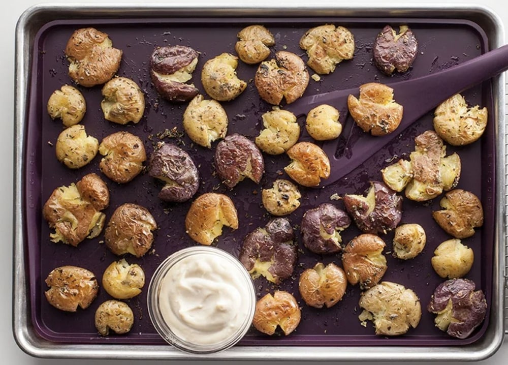 A sheet pan of crispy smashed potatoes with a bowl of creamy dip and a purple spatula
