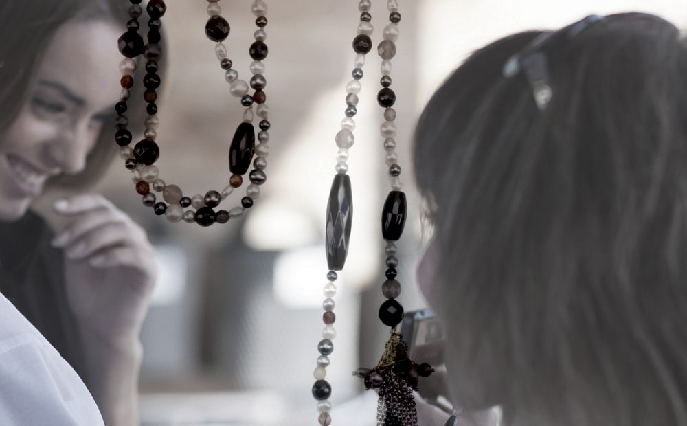 Two women smiling as they look at long beaded necklaces of dark stones, pearls and a tassel pendant
