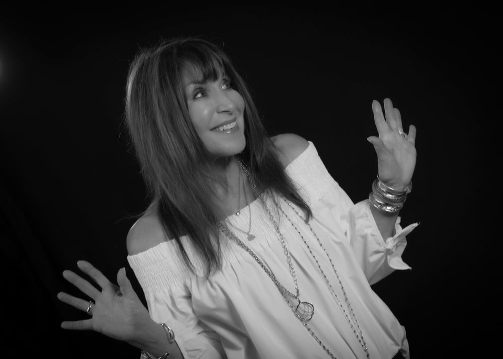 Black and white photo of designer Sara Blaine smiling with her hands raised, wearing layered necklaces and bangles