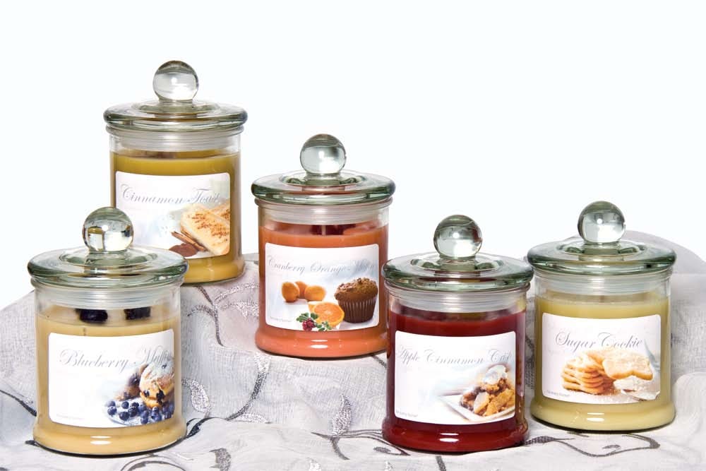 Five lidded glass jar candles in bakery scents such as cinnamon toast, cranberry orange muffin and sugar cookie on a draped tablecloth