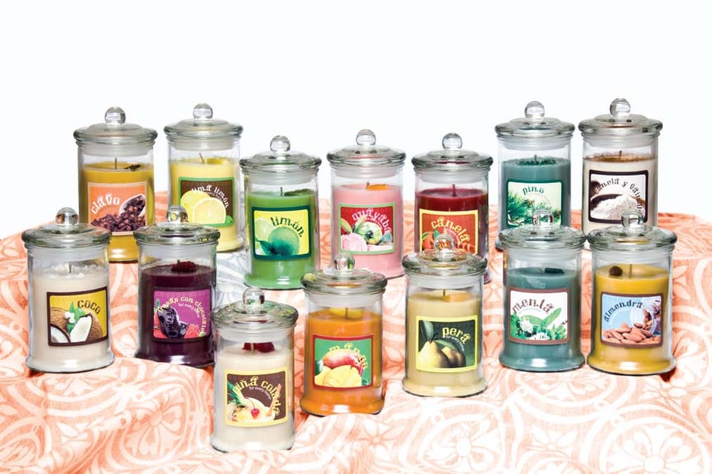 Fourteen lidded jar candles with Spanish-language fruit and spice labels such as coco, limón and pera on a peach patterned cloth