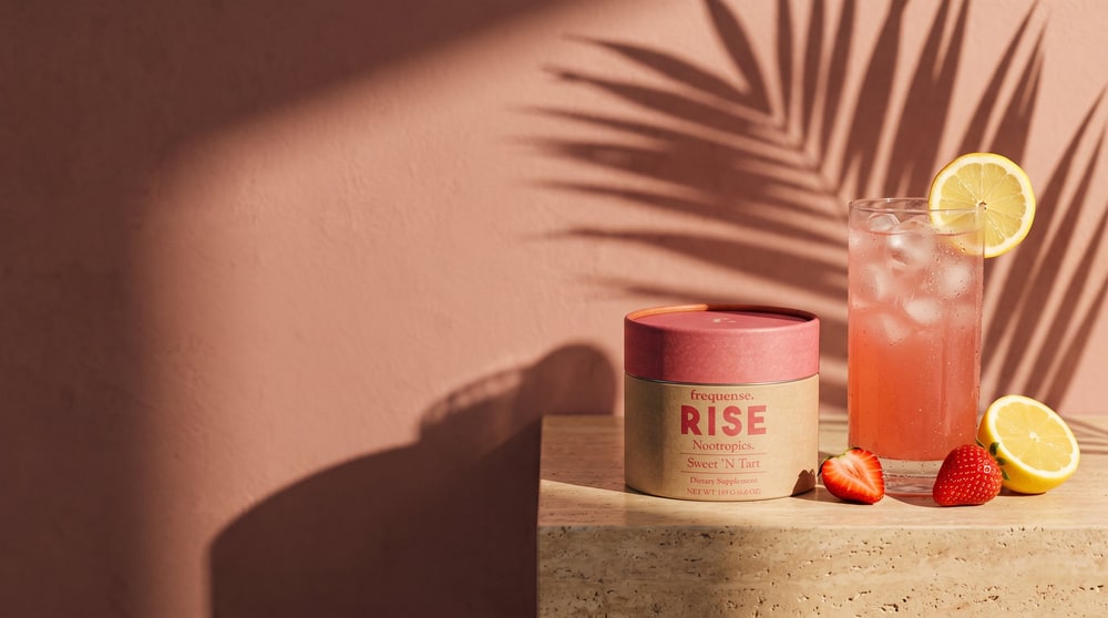 A glass of iced pink drink with lemon and strawberries beside a tub of Frequense RISE Sweet 'N Tart on a stone block, palm shadows on a terracotta wall