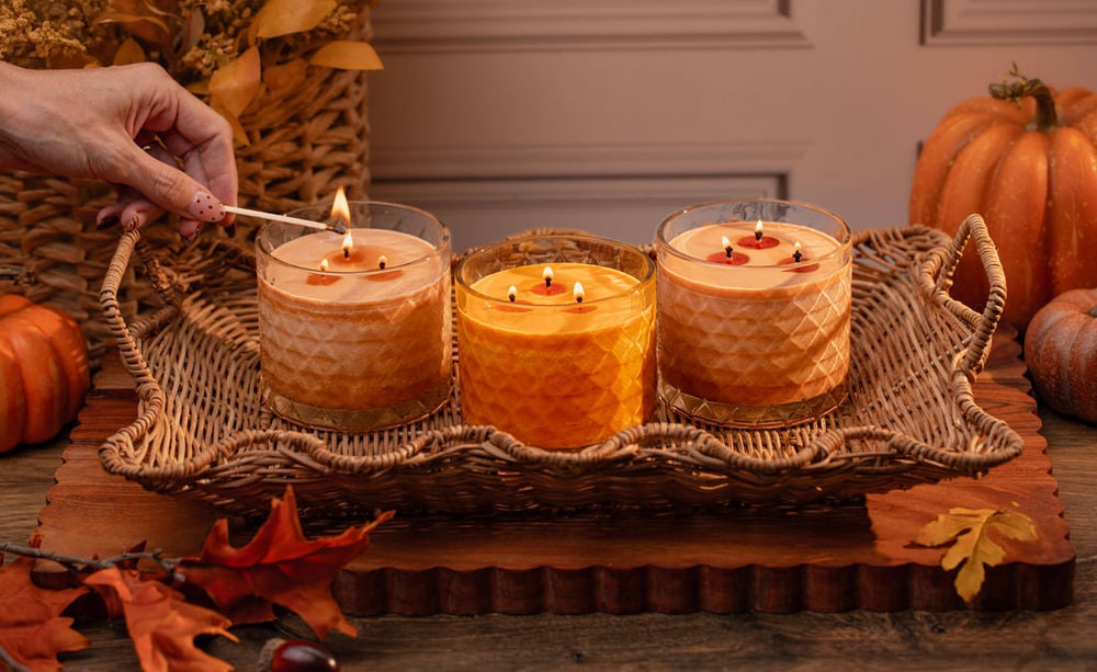 A hand lighting three Gold Canyon jar candles in amber and orange glass, set in a wicker tray surrounded by pumpkins and autumn leaves