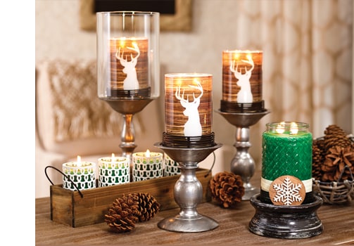 Gold Canyon candles in deer-silhouette glass holders on silver pedestals, with a green jar candle, small tealights and pine cones on a wooden table