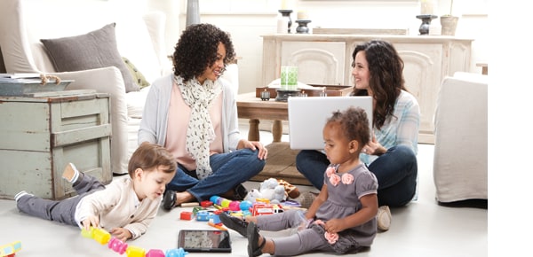 Two women sitting on a living room floor with a laptop while two toddlers play with toys beside them