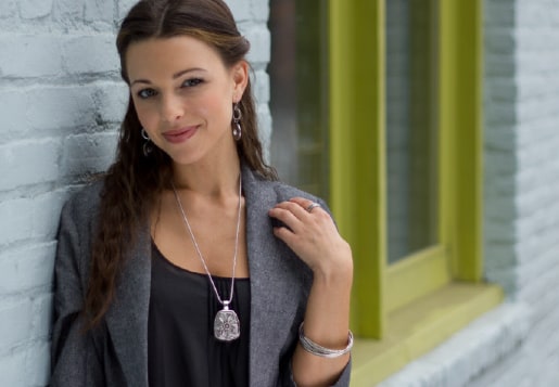 A woman in a grey jacket leaning against a white brick wall, wearing a long silver pendant necklace, drop earrings and a bangle