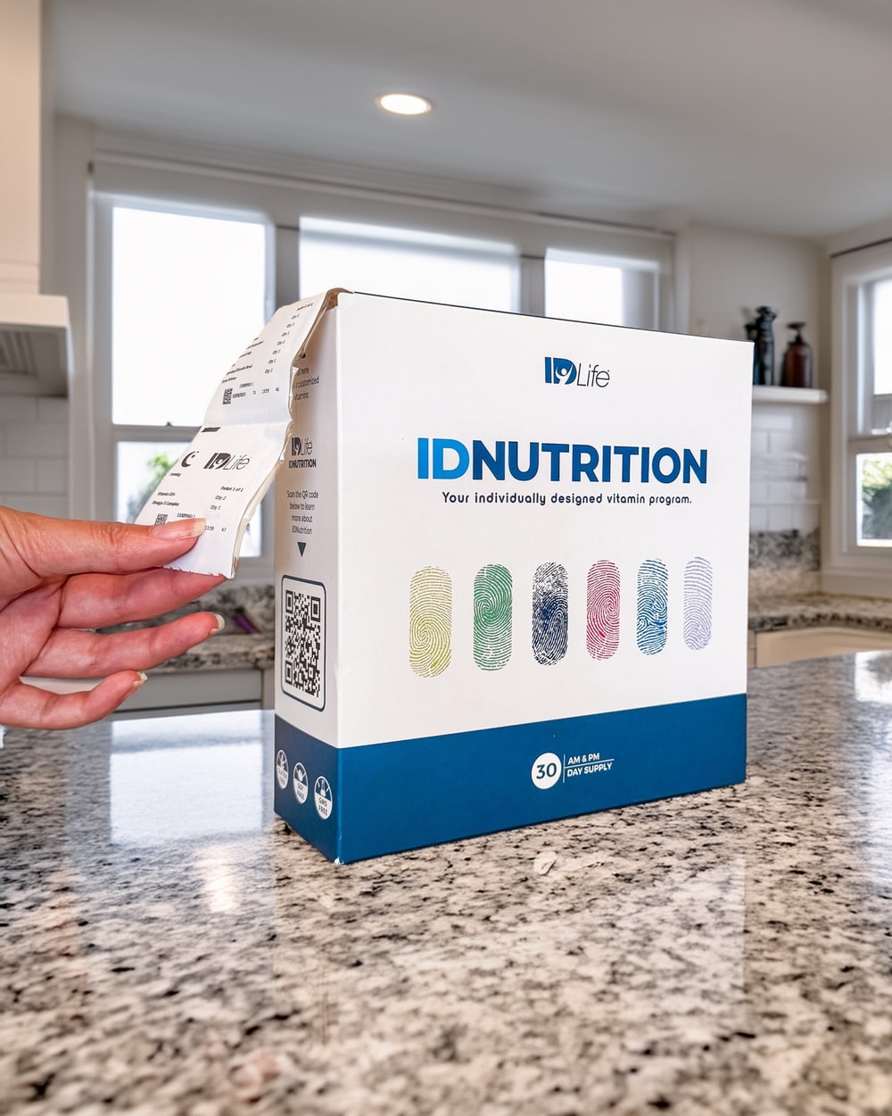 A hand pulls a strip of personalized vitamin packets from an IDNutrition box on a granite kitchen counter