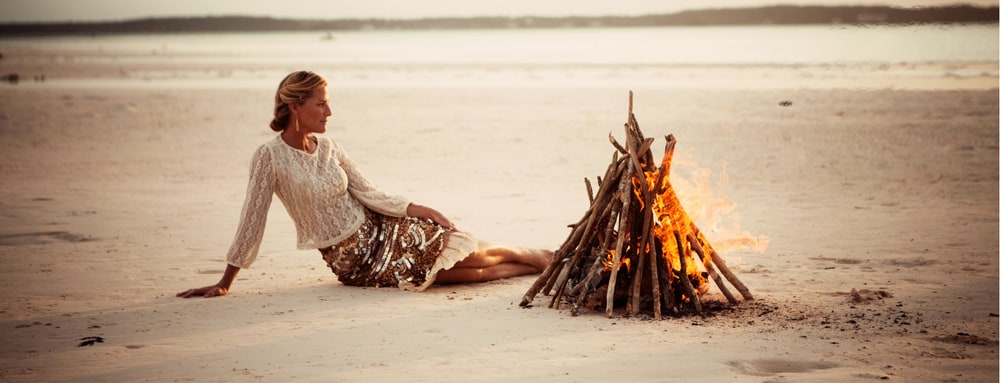 A woman in a cream knit sweater and sequined skirt sitting on a wide sandy beach beside a small bonfire at sunset