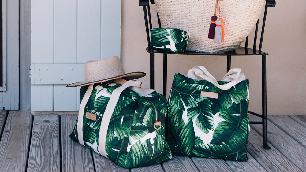 Green and white palm-print India Hicks travel totes on a wooden porch beside a chair holding a straw bag